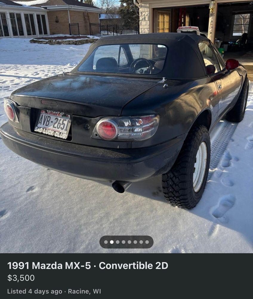 1991 Mazda Miata, lifted with big winter wheels/tire, what look like RX8 taillights, car painted black. 
