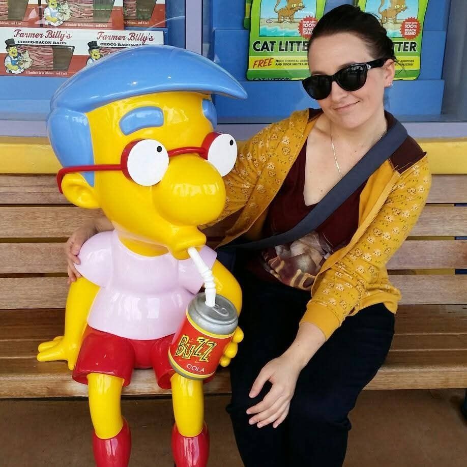 A photo of a white brunette woman in a yellow jacket, maroon tshirt and black jeans is sitting next to a fibreglass statue of Millhouse, a cartoon character from The Simpsons, on a bench. She is wearing sunglasses and her arm is around him. He is yellow, wears glasses and has blue hair, and is drinking a buzz cola.