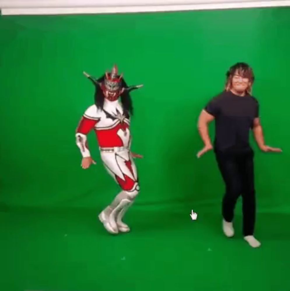 a man in a superhero costume is dancing next to another man in a black shirt in front of a green screen .