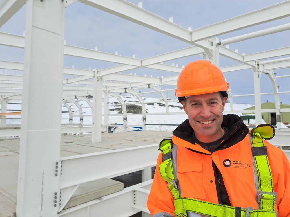 A smiling man wearing protective clothing for construction and polar weather stands next to a skeleton framework for a new building. There are snowy mountains in the background.