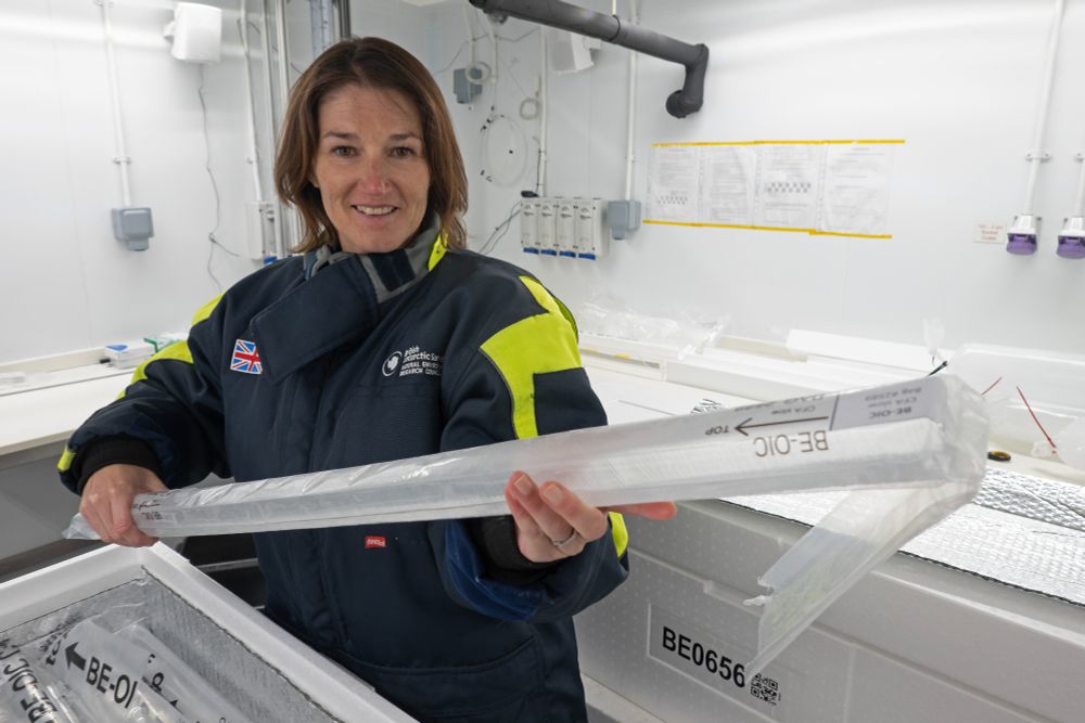 Liz stands in a walk-in scientific freezer, wearing a warm jacket. She's holding a long ice sample - a narrow stick of ice about a metre long and only a few centimetres wide. It's from a box containing many ice cores. Photo credit: Jamie Oliver, BAS.