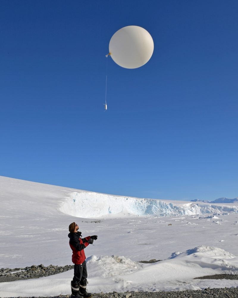 A person in winter gear releasing a large white weather balloon into a clear blue sky over a snowy, icy landscape.