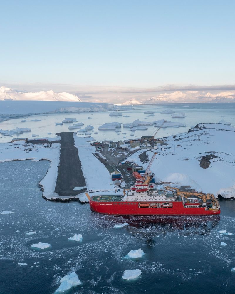 Red icebreaker ship the RSS Sir David Attenborough docked at a snowy Antarctic research station surrounded by floating icebergs and calm waters.