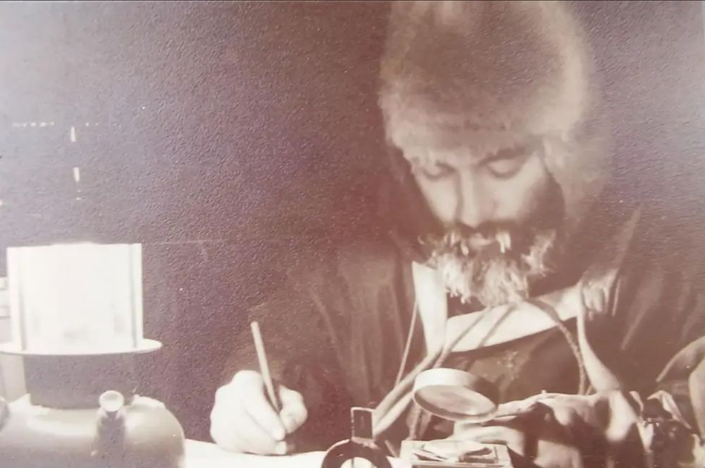Dennis Bell pictured during his 2-year assignment in Antarctica, shared by his brother David Bell. He is recording measurements by lamplight with a magnifying glass, dressed in a furry hat and with icicles in his moustache.