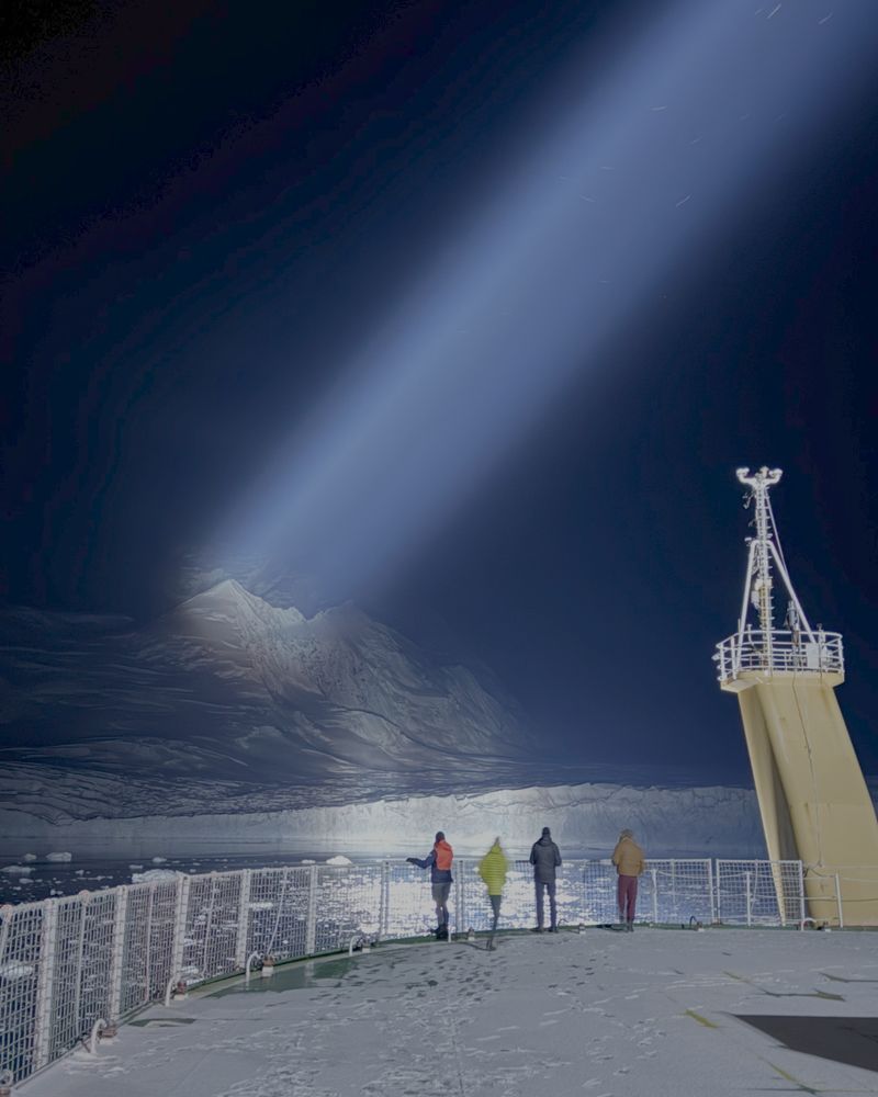Four people stand on the ship's helideck, looking at a glacier that is being lit up by the ship's searchlight

