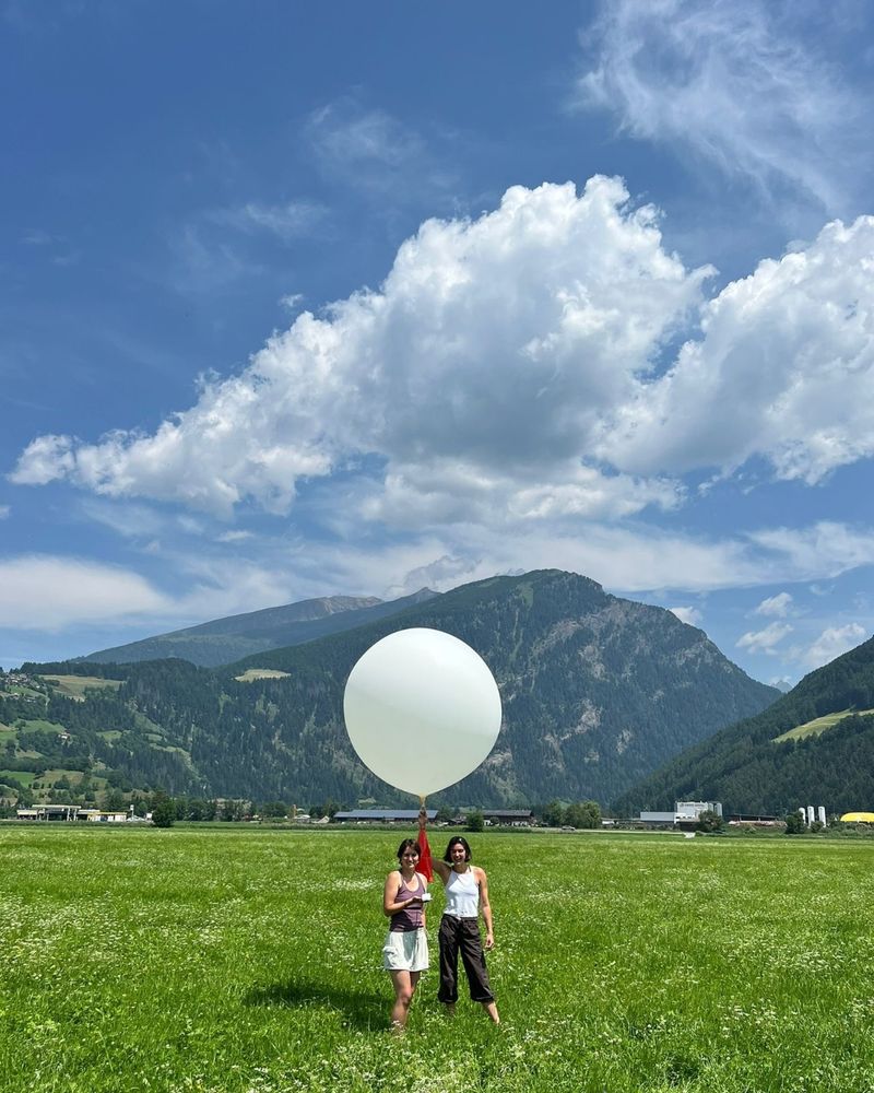 Two people standing in a green field holding a large white weather balloon under a partly cloudy blue sky with mountains in the background.