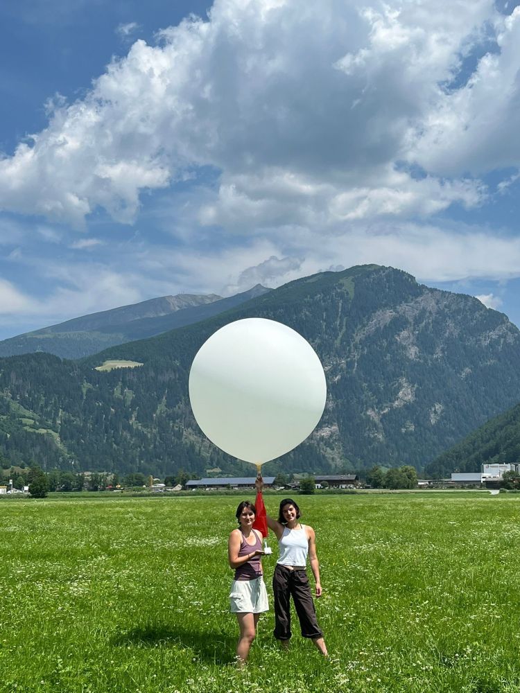 Two people standing in a green field holding a large white weather balloon under a partly cloudy blue sky with mountains in the background.
