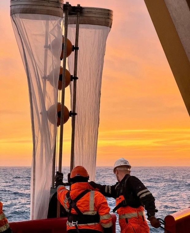 Two workers in orange safety gear operate large vertical Bongo nets on a boat at sunset over the ocean.