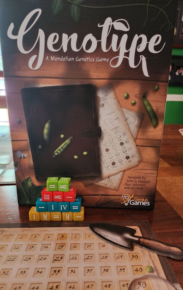 Cover box for the board game Genotype by Genius Games.  Box features a drawing of an old leather journal with splayed pages for listing genetic characteristics of pea plants.  Several drawn green pea pods are also shown on the box.  In front of the box is a pyramid of colored dice (green, red, blue, and orange) depicting roman numerals  on each side that are used in the game.  A score tracker is also shown on the table.