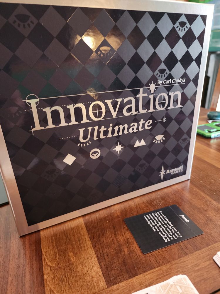 Cover box for board game Innovation Deluxe featuring a black diamond checkerboard pattern on the background.  Also shows 5 symbols (diamond, eye, Starburst, mountain, and sparkle) representing the 5 included expansions