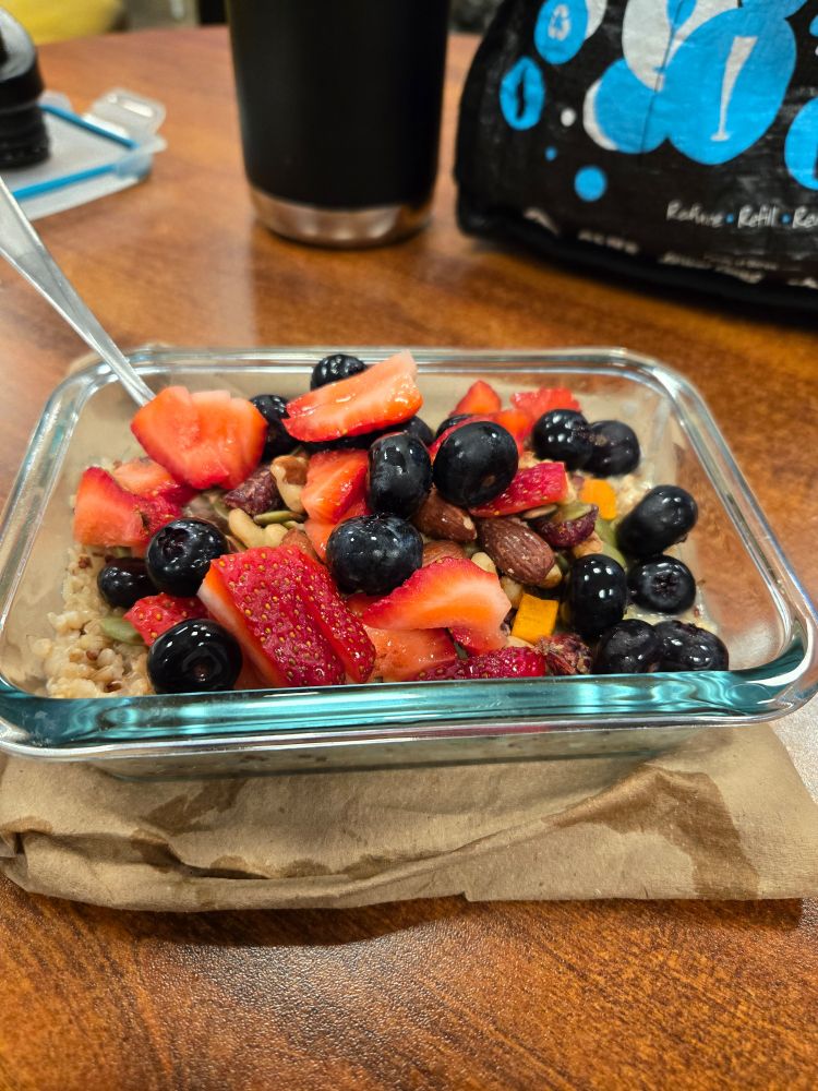 A glass dish with oatmeal on a table, topped with lots of strawberries, blueberries, and almondy trail mix. Real gourmet shit.