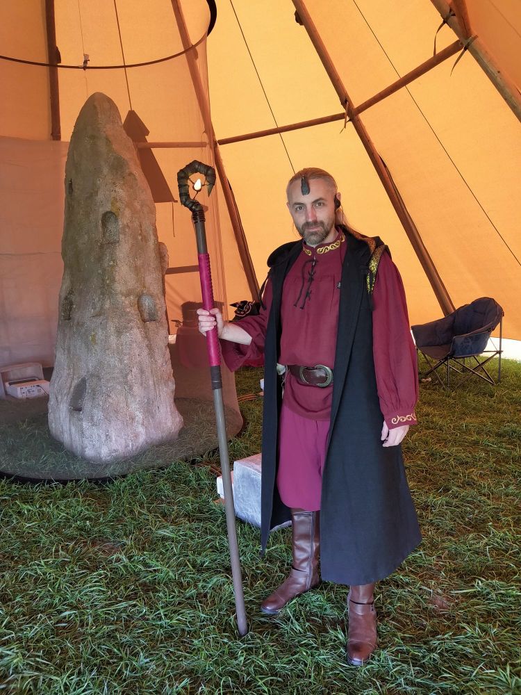 Chris, dressed as a wizard, standing in front of a standing stone inside a huge tent.