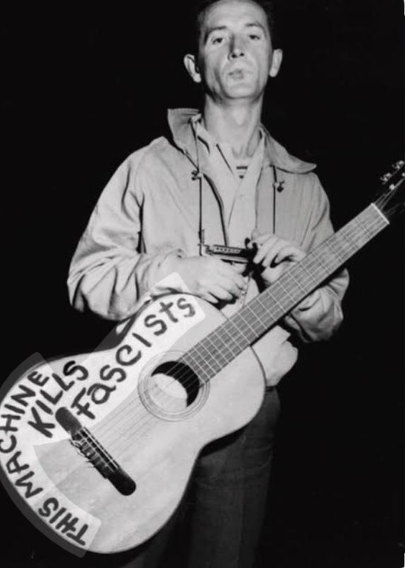 A man is standing with his guitar. On the instrument there is printed a text: "This machine kills fascists".