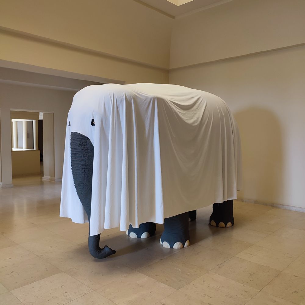 A large sculpture of an elephant covered with a plain white sheet, leaving only its trunk, feet, and parts of its ears visible. The sheet drapes heavily over the animal, creating folds that emphasize its hidden bulk. The scene is set indoors in a neutral-colored room with tiled flooring and beige walls. 