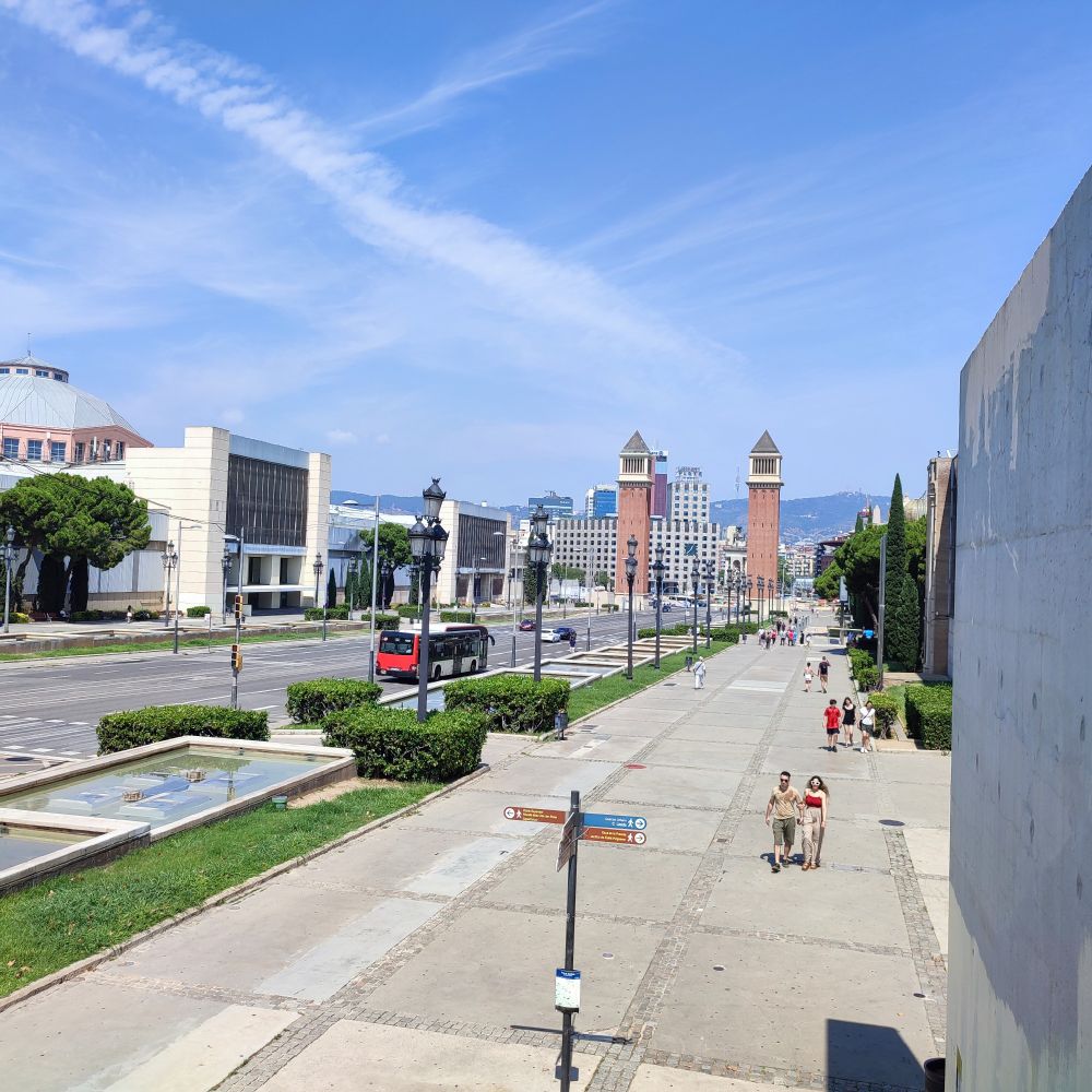 View down a wide pedestrian walkway and parallel avenue in Barcelona, with the Venetian Towers in the background, a red and black bus on the road, and several people walking under a sunny blue sky.