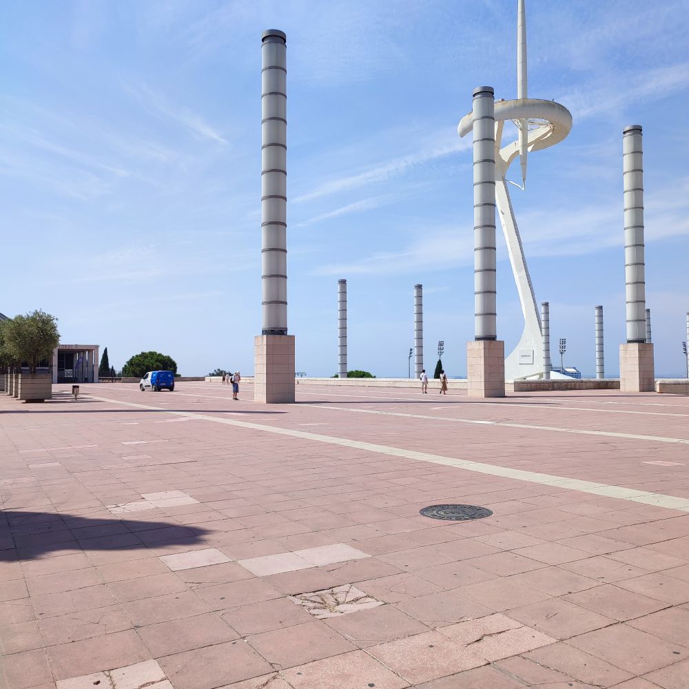 Open plaza with tall cylindrical columns and the white, futuristic Montjuïc Communications Tower in Barcelona, under a bright, partly cloudy sky