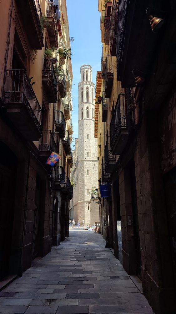 Narrow, shaded street lined with tall, closely spaced buildings and balconies, opening onto a sunlit square with a tall, octagonal bell tower of a stone church rising prominently against a clear blue sky.