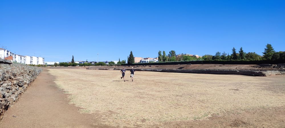 Two people are running side by side on the dry, flat ground of an ancient Roman circus ruin, with low stone walls visible on the left and right. In the background are modern buildings, trees, and a bright blue sky without clouds.