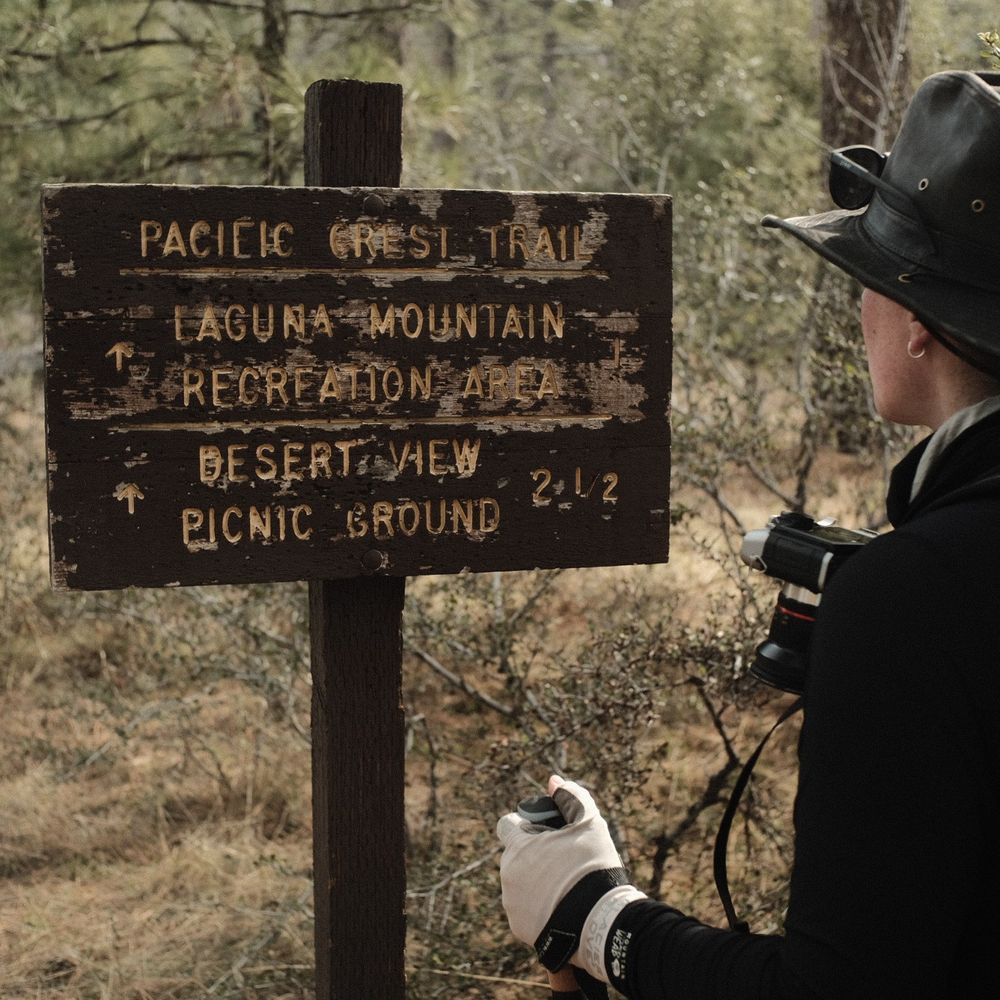 Kiri on the right of the frame, looking at a large, wooden sign with old, yellow lettering on it: "Pacific Crest Trail, Laguna mountain recreation area 1, desert view picnic ground 2 1/2"