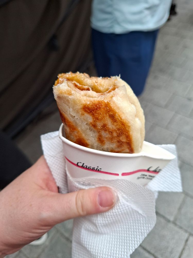 Ein gefalteter Hotteok in einem Pappbecher wird von einer Hand gehalten. Er ist angebissen.