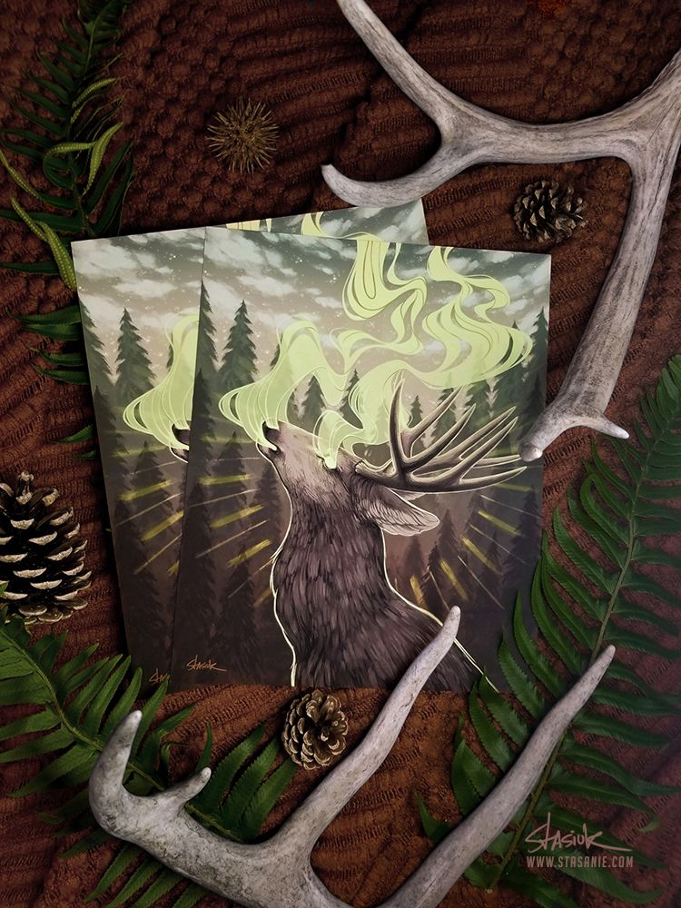 A photograph of 2 art prints on a dark blanket. The art prints are of the illustration "Release" by Stephanie Stasiuk Monk. The prints are surrounded by pinecones, ferns, and antlers.