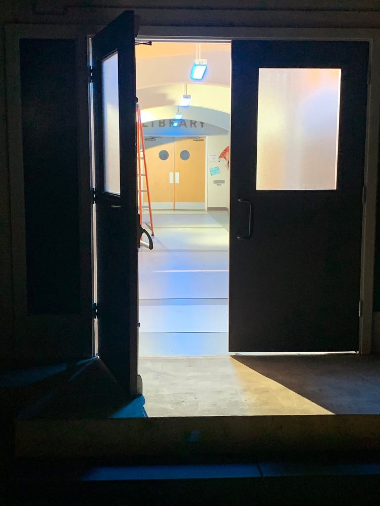 An open door below a sign with the same border as the old Sunnydale High sign. In the background of a school highway you see a set of swinging doors with small circular windows, above which the word LIBRARY is on the wall. 
