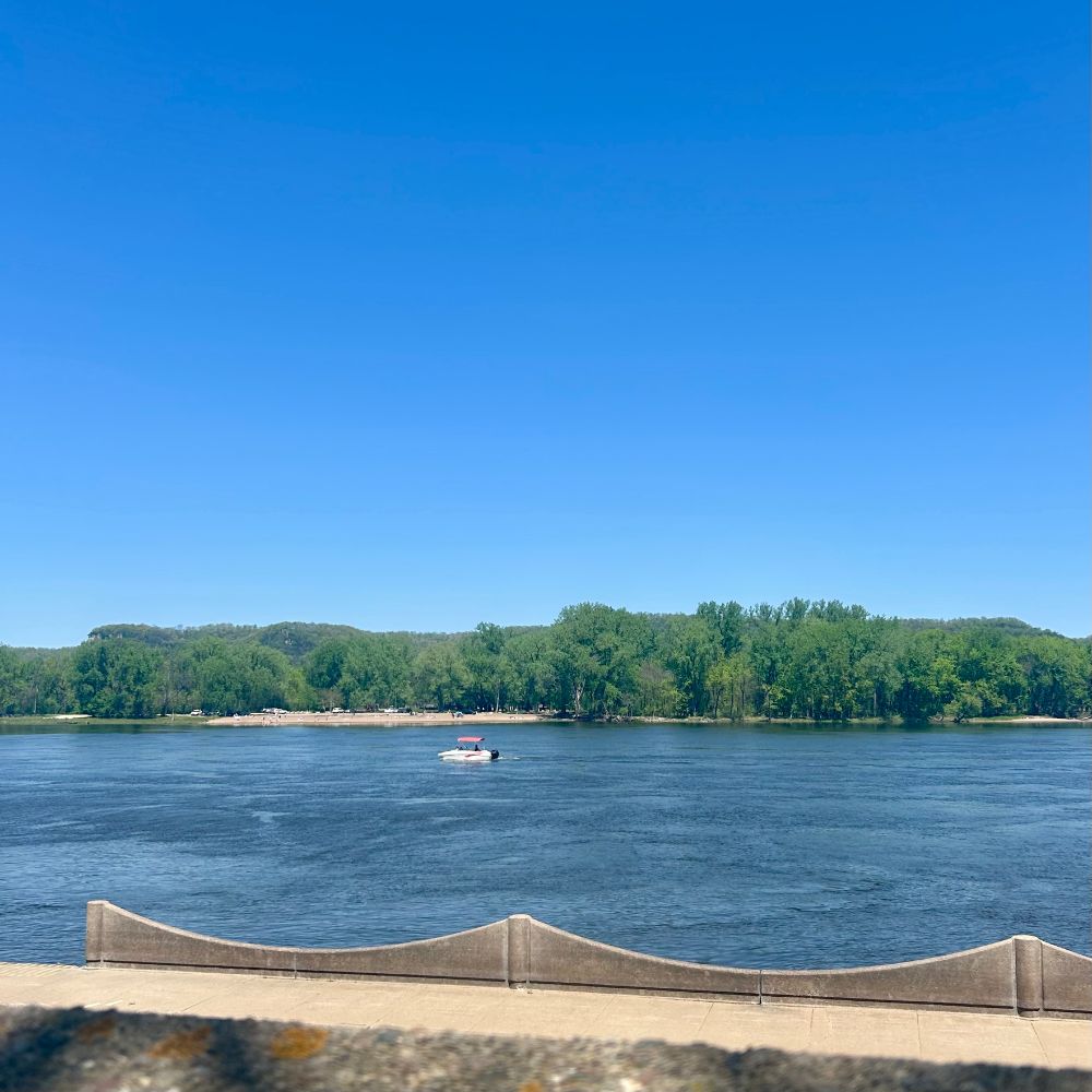 A view across the Mississippi River, in the distance is the far shore with sandy beach between green trees. In the middle of the river is a small boat moving upstream. The river and the sky are intensely blue.