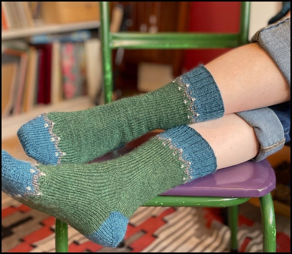 A photo of crossed legs resting on a green and purple chair with a bookshelf and red wall behind. The feet are in green socks with blue-green toes, heels, and cuffs.