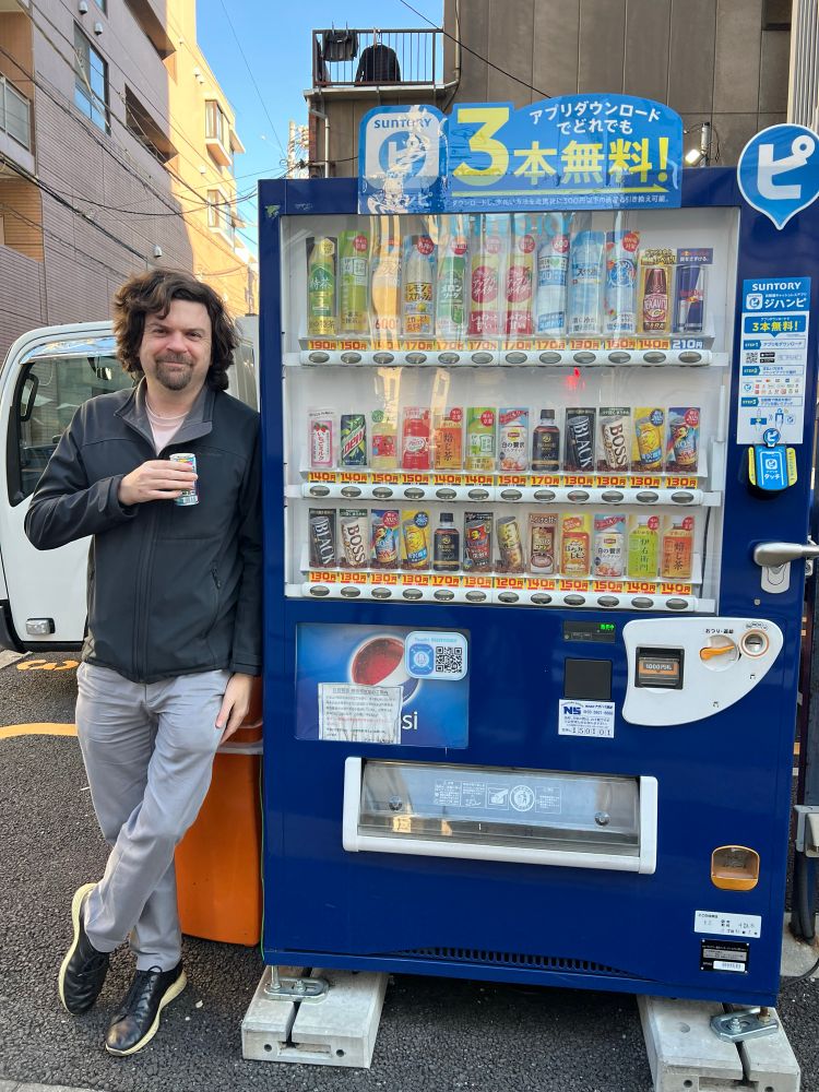 Brendan leaning against a large vending machine with 3 rows of assorted drinks. He is holding a small can of coffee. He looks like he needs it, and is happy to have found it