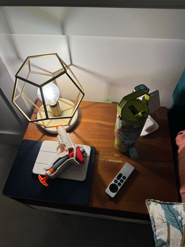 A nightstand table with a nerf gel blaster, iPad, laptop, remote, and water bottle. 