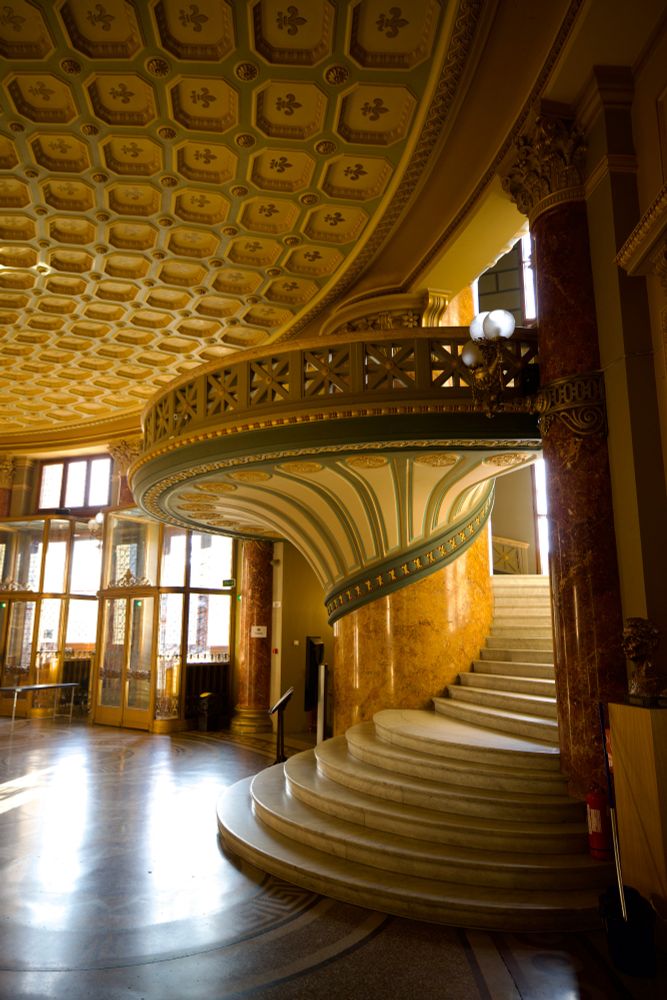 A spiral staircase cascades down an old theater atrium Soft yellow light illuminates the room.