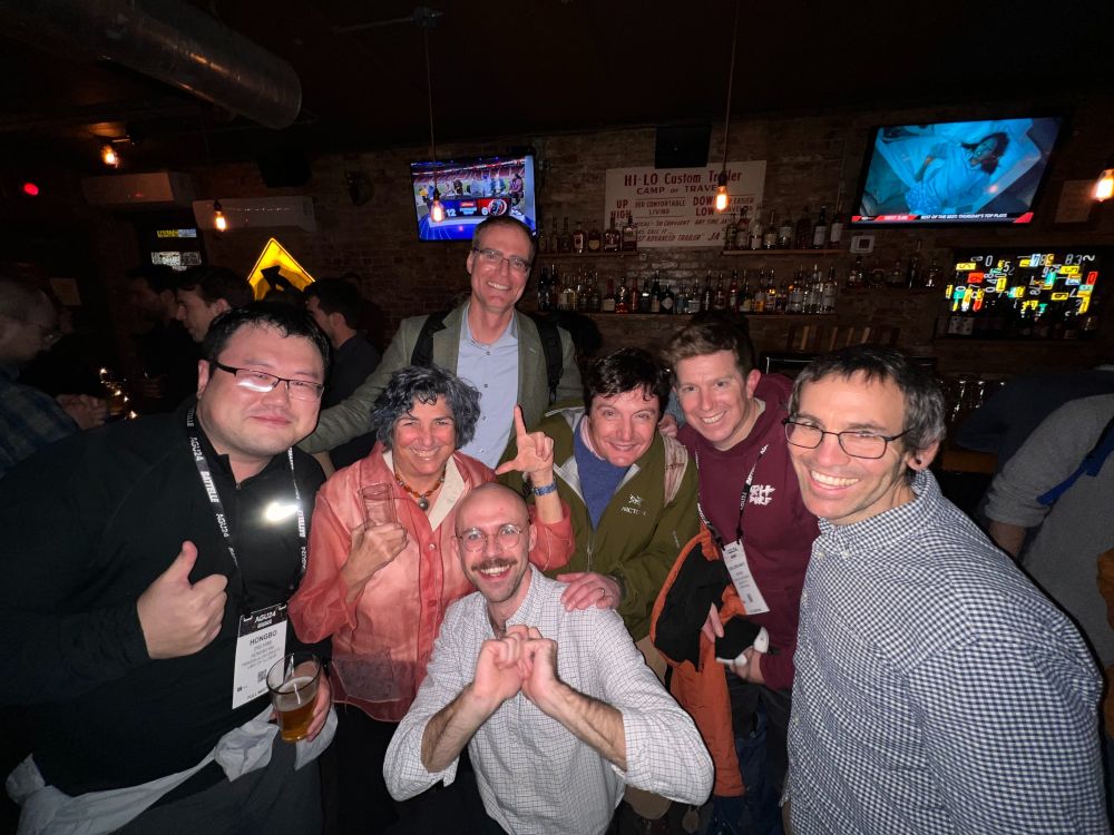 five men and one woman surrounding a man  -with many showing a thumbs up- in a bar setting