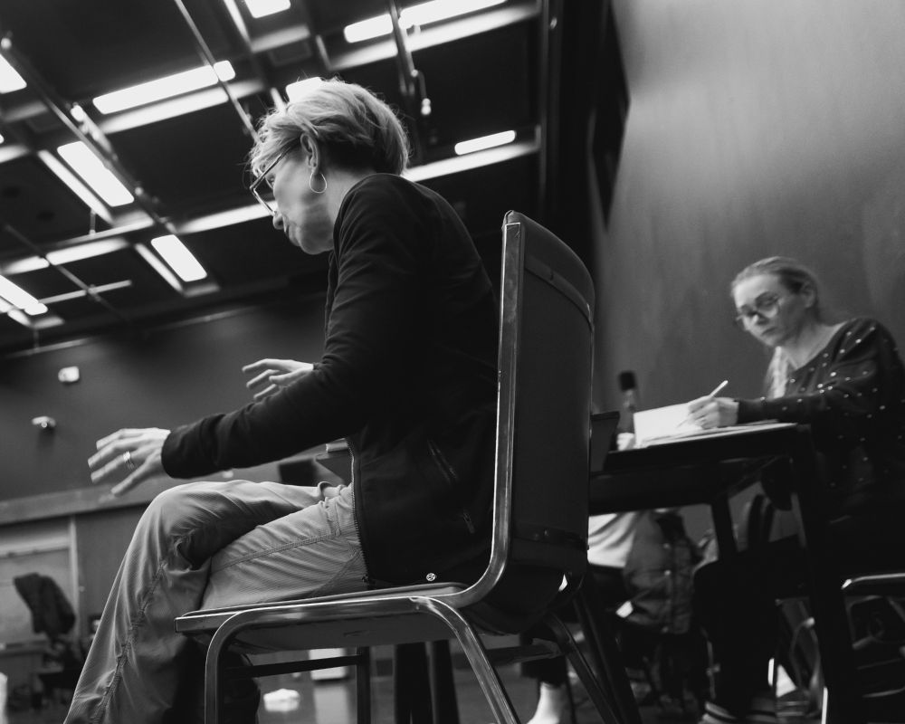 photo of a woman wearing glasses, gesturing with her hands, directing a play. to her right is another woman wearing glasses following the script and making notes. the room is an acting studio from the 1960's