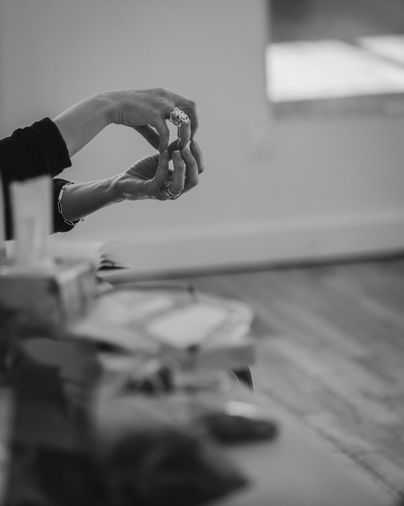 black and white photo of a woman's hands describing something. a silver ring on her upper hand. table in the foreground full of stuff