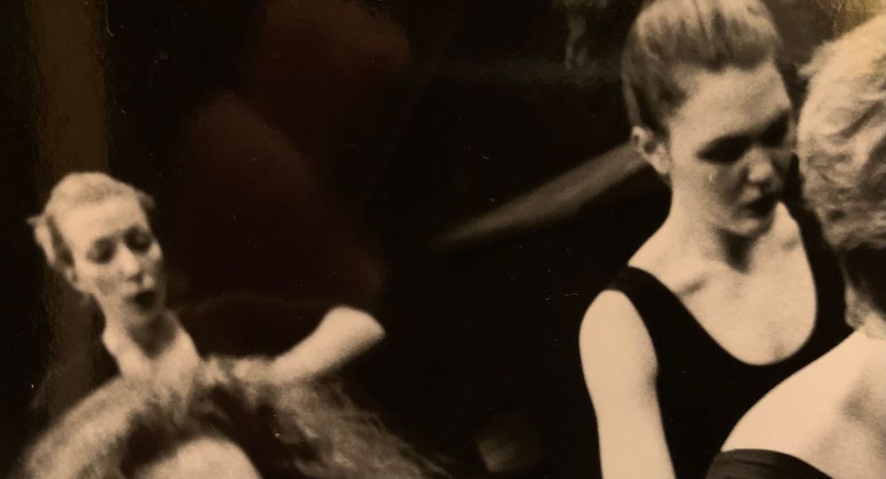 photo of two women. gaze down.  both hair pulled back. they're making some dance-theatre. photo of parts of two other women's heads in the foreground.