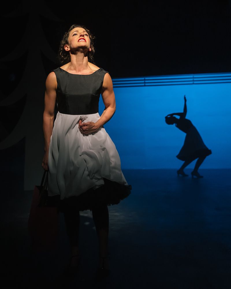 photo of a woman in a dress with a black top and white skirt in the foreground while a woman in the same dress plus a hat us upstage doing a back bend. blue lighting.