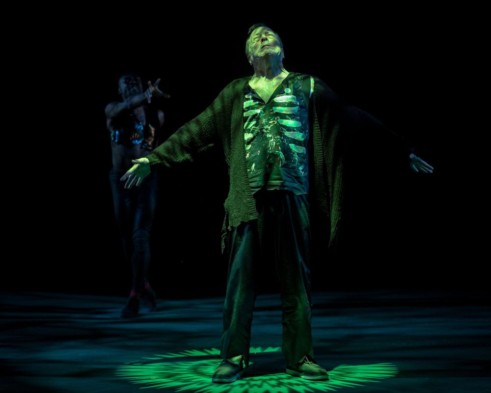 photo of a man on the right in the shadows casting a spell on a man standing in green light wearing a short with skeleton ribs on it and cardigan