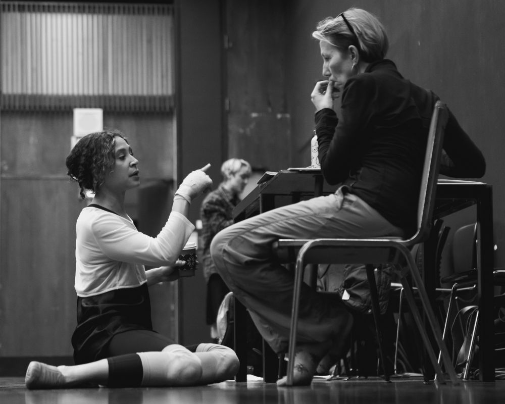 photo of an actor sitting on the floor talking to a director sitting in a chair chewing on her finger nails. a rehearsal room. an actor in the background on break.