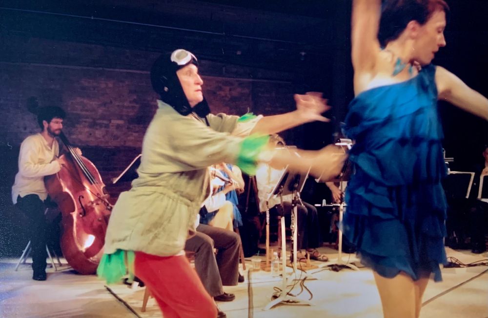 man in rustic shirt is playing a stand-up base while a woman in orange tights, aviator hat and green utility jacket (she's playing a duck) spins another woman in a royal blue flapper dress with yellow leggings (she is playing a bird. a small chamber orchestra (flute, violin etc) is obscured in the background)