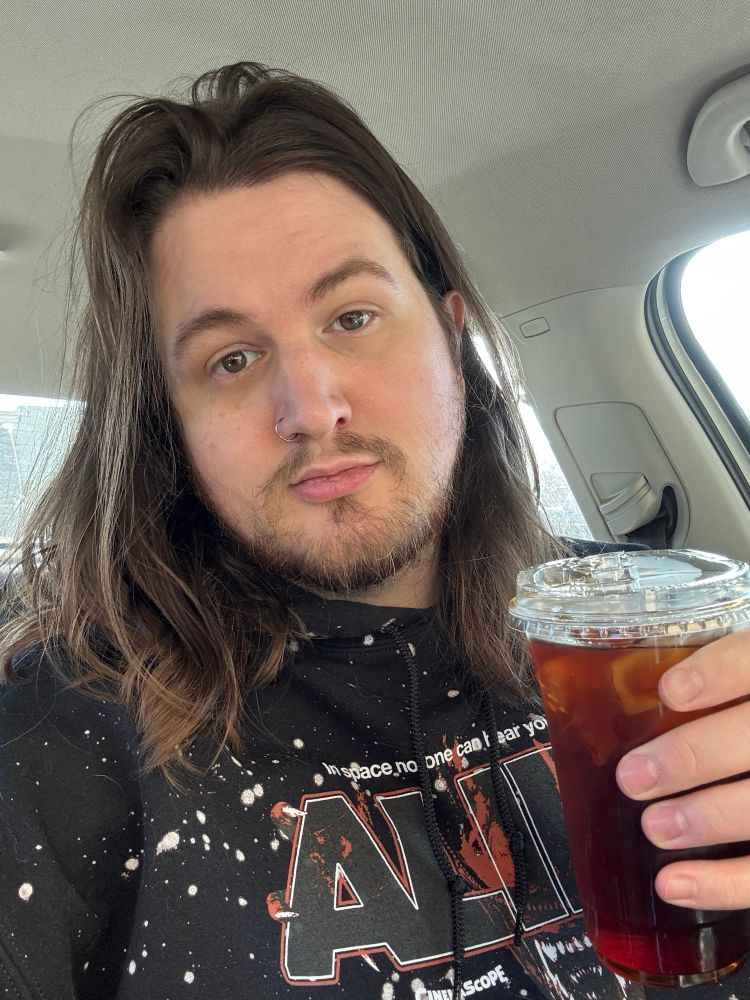 a man with shoulder length hair sitting in a car wearing an Alien hoodie and holding a very mid cup of cold brew