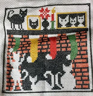 "Waiting for Santa" by The Stitcherhood. Silhouette of three white cats in front of a fireplace. One of the white cats is batting at a green stocking hung on the mantel. A black cat stands on the mantel next to a portrait of himself.  The mantel also has a photo of the three white cats. Stitched on 14ct white aida.