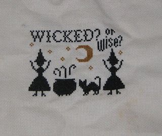 "Wicked" by the Stitcherhood. Silhouettes of two witches with their arms up and a cat, standing next to a steaming cauldron with dark gold stars and a crescent moon in the background. Writing across the top says "Wicked? or Wise". Cross stitch on 18ct tea-dyed aida.
