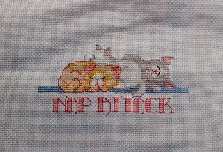 "Nap Attack" by Donna Kooler Designs. An orange, white and gray cat are sleeping in a pile over the words "Nap Attack" in pink. Cross stitched on 18 ct white Aida.