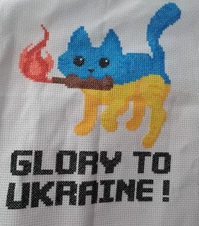 A blue and yellow cat holds a molotov cocktail. "Glory to Ukraine!" in block letters across the bottom. Cross stitch on 14ct white aida. Design by CuteFoxyShop of Poltava.