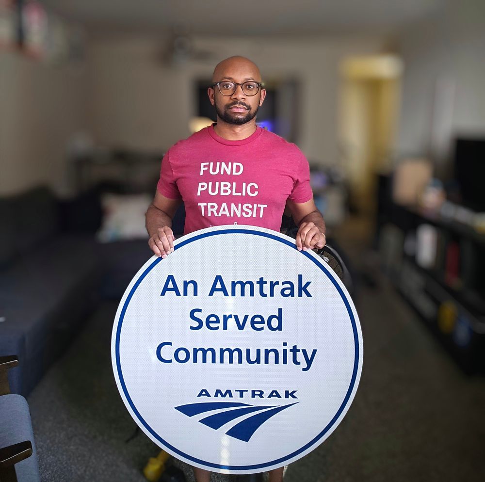 Me wearing a Fund Public Transit shirt while holding a sign that reads "An Amtrak Served Community." 