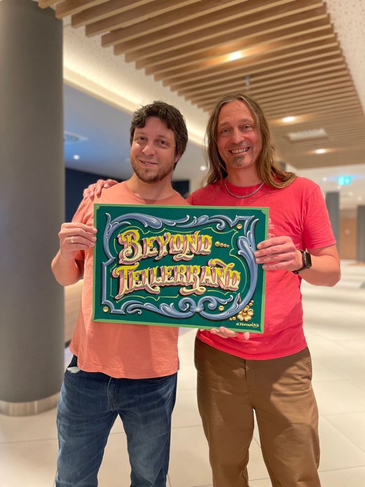 A photo that show Gustavo handing over the finished painting to Marc. Both holding the sign into the camera together smiling happily.