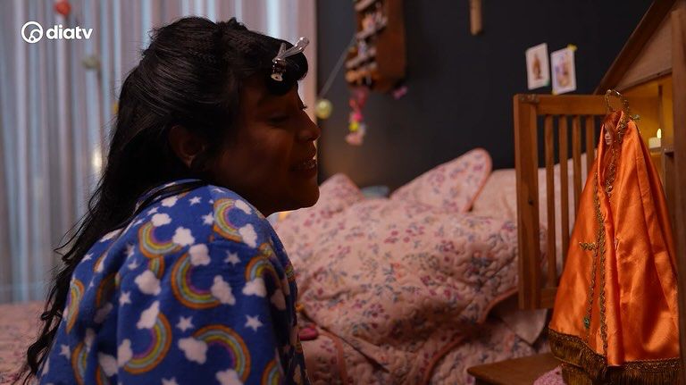 Cena da novela Blogueirinha, a Feia, em que a protagonista Blogueirinha está ajoelhada diante da imagem da Santa Mari Maria.