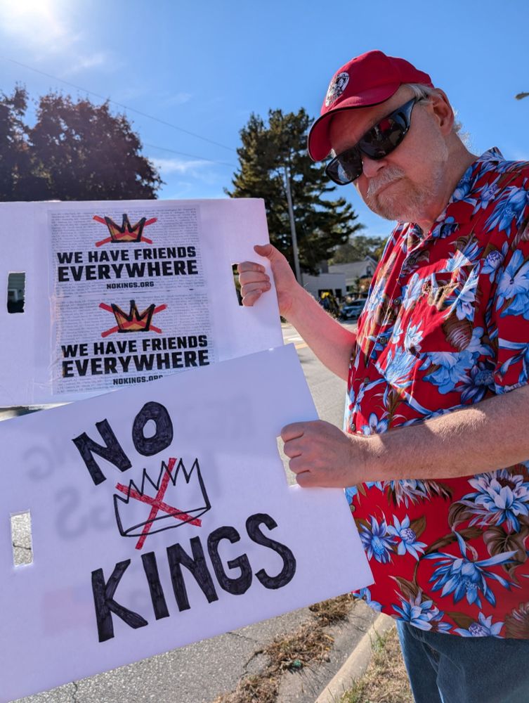 Big scruffy dude in bright colors and sunglasses holding two signs, one saying We Have Friends Everywhere and No Kings 