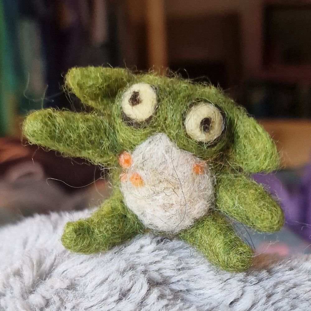 A frog goblin made of felt. He has oogly eyes and big ears