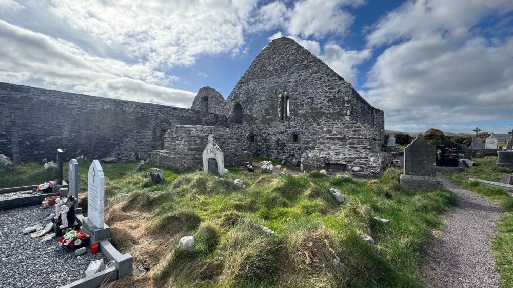 The ruin of Ballinskelligs Abbey. Cloudy sky in the background. Graveyard mostly overgrown with tall grass in the foreground.